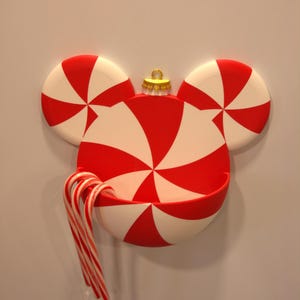 May include: A red and white peppermint-patterned wall-mounted holder shaped like a mouse head with circular ears. The holder features a gold-colored ornament cap at the top and contains three red and white striped candy canes hanging over the front edge.