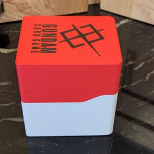 May include: A red and white card game box. The top section is red with the words "GUNDAM CARD GAME" and a black graphic. The bottom section is white. The box is rectangular and likely made of plastic.