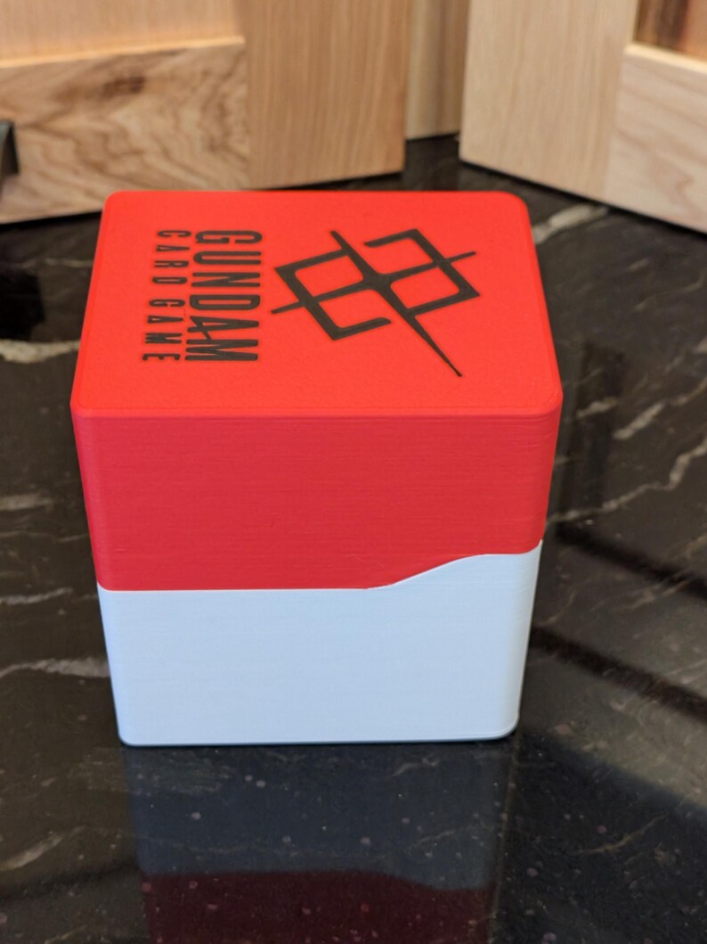 May include: A red and white card game box. The top section is red with the words "GUNDAM CARD GAME" and a black graphic. The bottom section is white. The box is rectangular and likely made of plastic.
