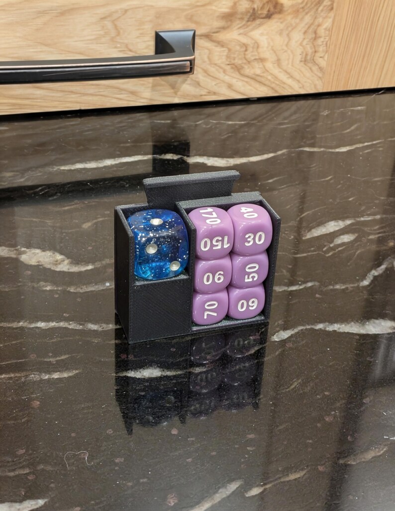 Puede incluir: Un soporte para dados negro impreso en 3D con dos compartimentos. Uno contiene un dado azul lleno de purpurina. El otro contiene seis dados morados con n&uacute;meros blancos. Los dados est&aacute;n numerados 06, 09, 30, 40, 50, 70, 150 y 170. El soporte est&aacute; sobre una superficie negra brillante.