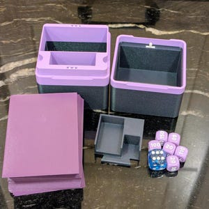 Puede incluir: Un conjunto de accesorios de juego, que incluye cajas de almacenamiento bicolores moradas y negras, una pila de fundas de cartas moradas, una peque&ntilde;a bandeja gris y un juego de dados morados y azules. Sobre una superficie oscura y estampada.