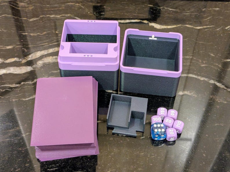 Puede incluir: Un conjunto de accesorios de juego, que incluye cajas de almacenamiento bicolores moradas y negras, una pila de fundas de cartas moradas, una peque&ntilde;a bandeja gris y un juego de dados morados y azules. Sobre una superficie oscura y estampada.
