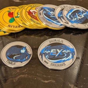May include: A selection of circular game tokens. Some are gold with character illustrations, others are silver with blue and black designs. The tokens feature text such as "MEGA EX HEADS" and "TAILS".