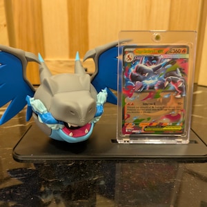 Puede incluir: Una figura de dragón gris y azul con la boca muy abierta y alas azules, junto a una carta de Pokémon en una funda protectora. La carta presenta una ilustración de dragón con el texto "Mega Charizard EX".