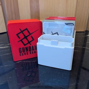 May include: A red card storage box with the text "GUNDAM CARD GAME" in white, next to a white card holder containing cards. The red box has a black graphic design. The card holder is open, revealing the cards inside.