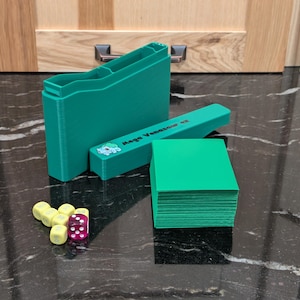 May include: A teal card game storage set with a matching deck of cards and dice. The set includes a card holder, a dice holder, and a deck of cards. The dice are yellow and purple. The card holder has the text "Mega Venusaur GX".