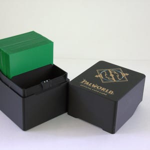 Könnte beinhalten: Eine schwarze Kunststoff-Deckbox für Sammelkarten mit abnehmbarem Deckel. Auf dem Deckel befindet sich der goldene Schriftzug Palworld Official Card Game sowie ein Logo. Im offenen Unterteil ist ein Stapel grünrückiger Karten zu sehen.