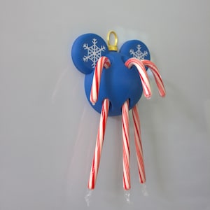 May include: A blue, spherical, mouse-shaped ornament featuring two circular ears with white snowflake patterns. The ornament has multiple holes containing red and white striped candy canes. A small gold loop is attached to the top for hanging.