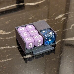 Puede incluir: Un soporte para dados gris oscuro con un compartimento para siete dados morados y un dado azul moteado. Los dados est&aacute;n numerados y el soporte est&aacute; sobre una superficie oscura.
