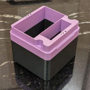 Puede incluir: Un contenedor de almacenamiento de dos tonos con una base negra y una parte superior lila. La secci&oacute;n superior tiene m&uacute;ltiples compartimentos y recortes. El contenedor est&aacute; hecho de un material impreso en 3D y se encuentra sobre una superficie reflectante.