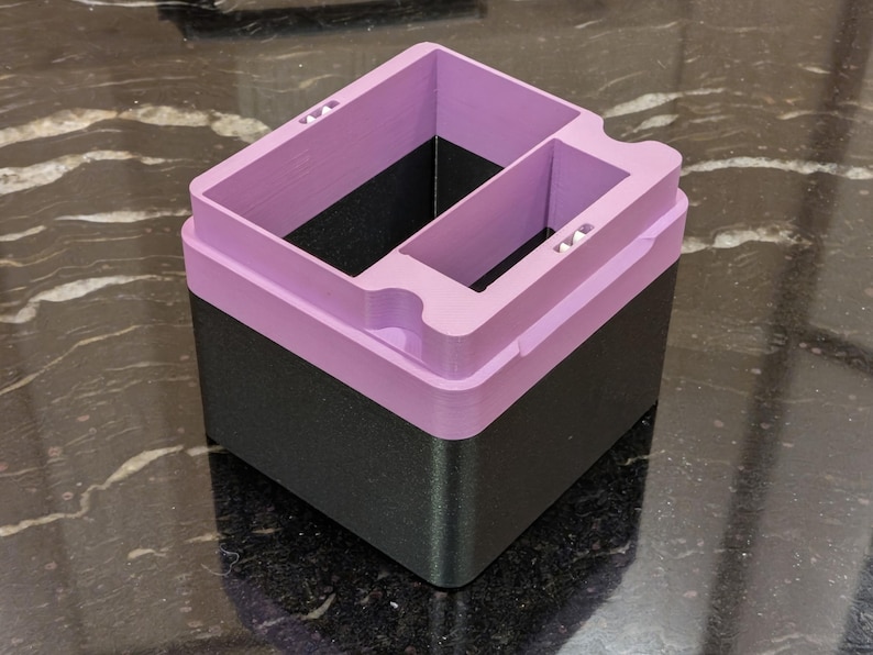Puede incluir: Un contenedor de almacenamiento de dos tonos con una base negra y una parte superior lila. La secci&oacute;n superior tiene m&uacute;ltiples compartimentos y recortes. El contenedor est&aacute; hecho de un material impreso en 3D y se encuentra sobre una superficie reflectante.