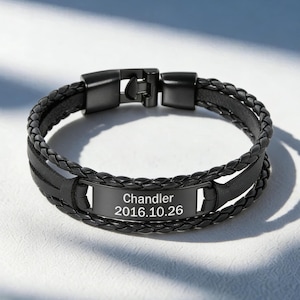 May include: Black leather bracelet with a braided design and a rectangular plate. The plate is engraved with the name "Chandler" and the date "2016.10.26". The bracelet has a clasp closure.