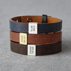 Puede incluir: Tres pulseras de cuero en azul marino, marrón y marrón oscuro, cada una con un acento metálico rectangular grabado con números romanos. Las pulseras están apiladas, mostrando los diferentes colores y texturas.