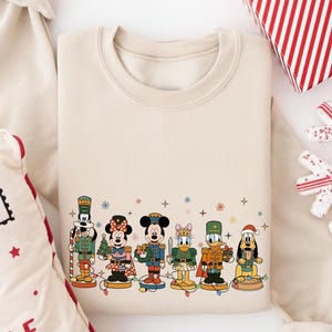 May include: Cream-colored sweatshirt featuring a festive design of Disney characters dressed as nutcrackers. The graphic includes Mickey, Minnie, Donald, and Goofy, with colorful accents and snowflake details. A holiday-themed gift is visible in the background.