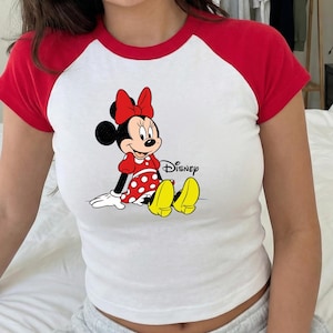 Puede incluir: Camiseta ragl&aacute;n blanca con mangas rojas con un gr&aacute;fico de Minnie Mouse. El personaje de dibujos animados lleva un vestido rojo con lunares blancos, un lazo rojo y zapatos amarillos. La palabra "Disney" est&aacute; impresa en negro.