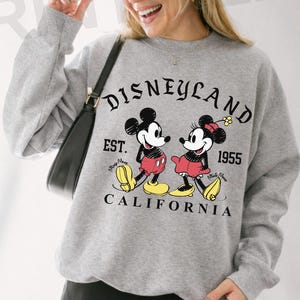 May include: Sport gray sweatshirt featuring Mickey and Minnie Mouse with the text "Disneyland Est. 1955 California". The graphic shows Mickey and Minnie Mouse in their classic outfits.