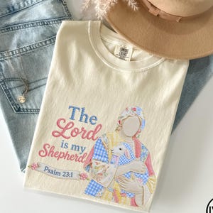 May include: Ivory-colored t-shirt with the embroidered text "The Lord is my Shepherd" and a graphic of a person holding a lamb. The shirt is paired with light blue jeans and a tan hat. The text "Psalm 23:1" is also embroidered on the shirt.