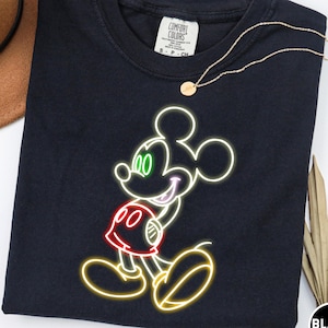 Op de afbeelding: Zwart T-shirt met een neon-stijl Mickey Mouse-ontwerp. De afbeelding bevat groene ogen, een rode broek en een gele omtrek. Het shirt heeft een ronde hals en een klein label op de bovenrug. Een gouden ketting is over het shirt gedrapeerd.