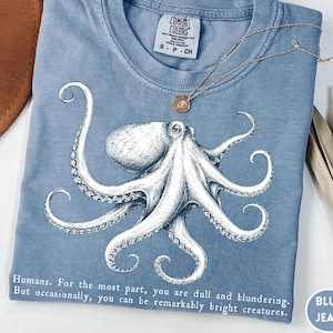 May include: A blue jean Comfort Colors t-shirt featuring a detailed white octopus illustration. The shirt includes the text: "Humans. For the most part, you are dull and blundering. But occasionally, you can be remarkably bright creatures."