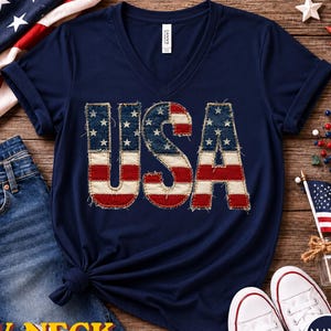 May include: Navy blue V-neck t-shirt with the word "USA" in a patriotic design. The letters are made to look like the American flag. The shirt is paired with blue jeans and white sneakers.