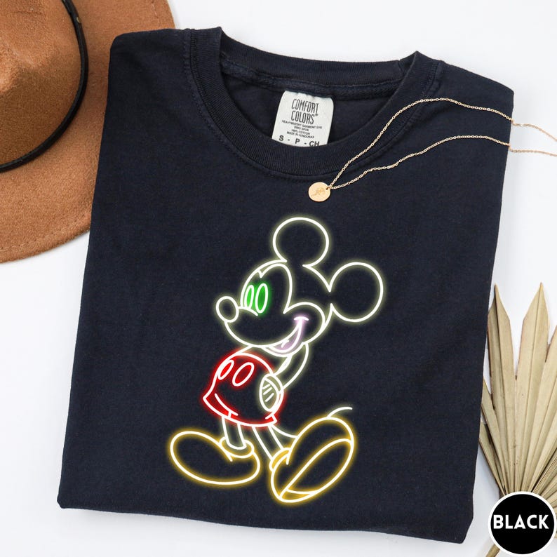 Op de afbeelding: Zwart T-shirt met een neon-stijl Mickey Mouse-ontwerp. De afbeelding bevat groene ogen, een rode broek en een gele omtrek. Het shirt heeft een ronde hals en een klein label op de bovenrug. Een gouden ketting is over het shirt gedrapeerd.