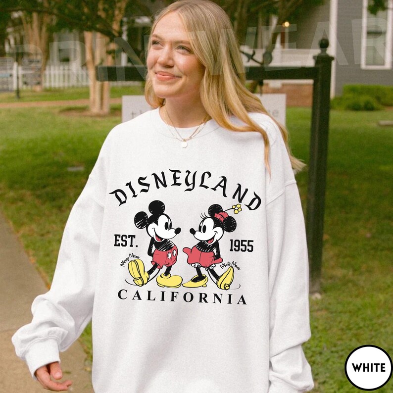 May include: White sweatshirt featuring a graphic of Mickey and Minnie Mouse with the text "Disneyland Est. 1955 California". The design includes Mickey in red shorts and Minnie in a red skirt. The sweatshirt has a crew neck.