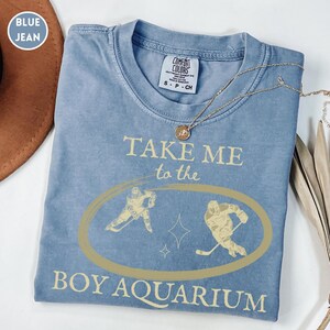 May include: A blue jean Comfort Colors t-shirt with the text "TAKE ME to the BOY AQUARIUM" in a light tan color. The shirt features a gold oval with two hockey players and a star design. A gold necklace is draped across the shirt.