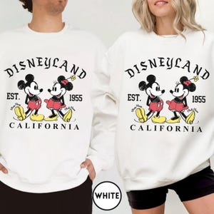 May include: White crewneck sweatshirt featuring a Disneyland graphic with Mickey and Minnie Mouse. The design includes the text "Disneyland," "Est. 1955," and "California." The cartoon characters are in red, black, yellow, and white.