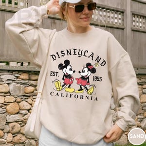 May include: Sand-colored Disneyland sweatshirt featuring Mickey and Minnie Mouse. The graphic includes the text "Disneyland Est. 1955 California". The long-sleeved crewneck sweatshirt is a casual style.