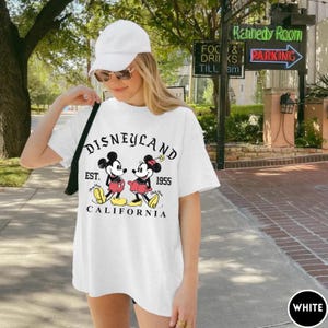 May include: White t-shirt with a Disneyland graphic featuring Mickey and Minnie Mouse. The design includes the text "DISNEYLAND EST. 1955 CALIFORNIA". The shirt is a classic, casual style.