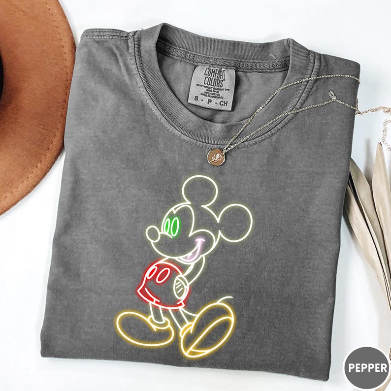 Op de afbeelding: Een grijs Comfort Colors T-shirt met een Mickey Mouse-ontwerp in neonstijl. De afbeelding bevat groene ogen, een rode broek en een gele omtrek. Een zilveren ketting met een hanger is over het shirt gedrapeerd.