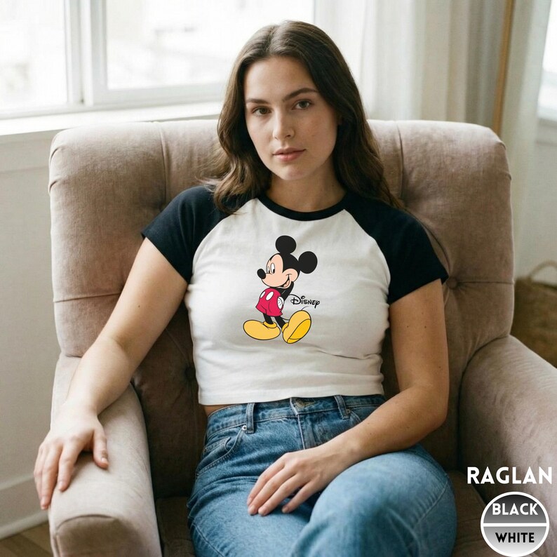 Puede incluir: Top ragl&aacute;n blanco y negro con un gr&aacute;fico de Mickey Mouse. El personaje de Disney se representa con pantalones cortos rojos, zapatos amarillos y orejas negras. El top se combina con vaqueros azules.