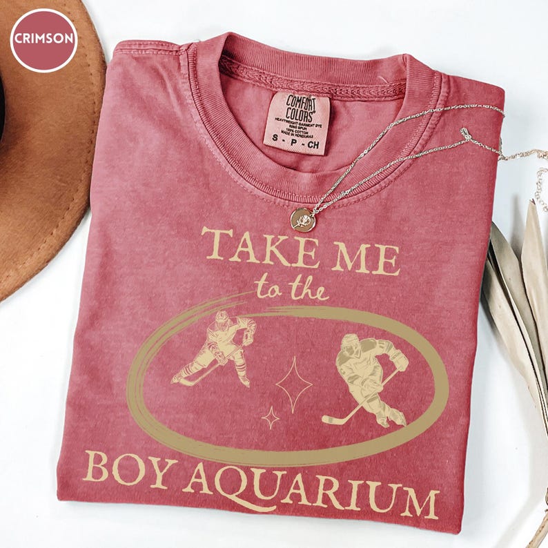 May include: Crimson-colored t-shirt with the text "TAKE ME to the BOY AQUARIUM" in a light tan color. The shirt features a graphic of two hockey players inside an oval. A silver necklace with a pendant is draped across the shirt.