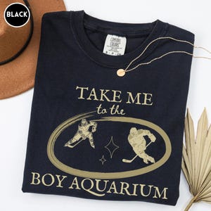 May include: Black t-shirt with gold text and graphics. The shirt reads "TAKE ME to the BOY AQUARIUM" with hockey player illustrations inside an oval. A gold necklace and a brown hat are also visible.