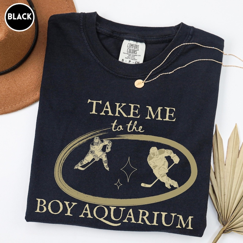 May include: Black t-shirt with gold text and graphics. The shirt reads "TAKE ME to the BOY AQUARIUM" with hockey player illustrations inside an oval. A gold necklace and a brown hat are also visible.