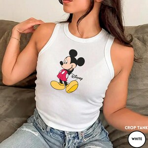 Puede incluir: Camiseta corta blanca con un gr&aacute;fico de Mickey Mouse. El personaje de dibujos animados lleva pantalones cortos rojos, zapatos amarillos y tiene orejas negras. La palabra "Disney" est&aacute; impresa debajo en escritura negra.