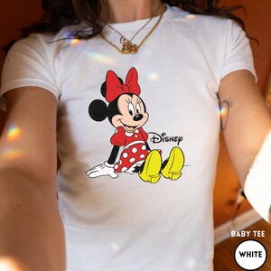 Puede incluir: Camiseta blanca para beb&eacute; con un gr&aacute;fico de Minnie Mouse sentada. Minnie Mouse lleva un vestido rojo con lunares blancos, un lazo rojo y zapatos amarillos. El logotipo de Disney est&aacute; impreso en negro.