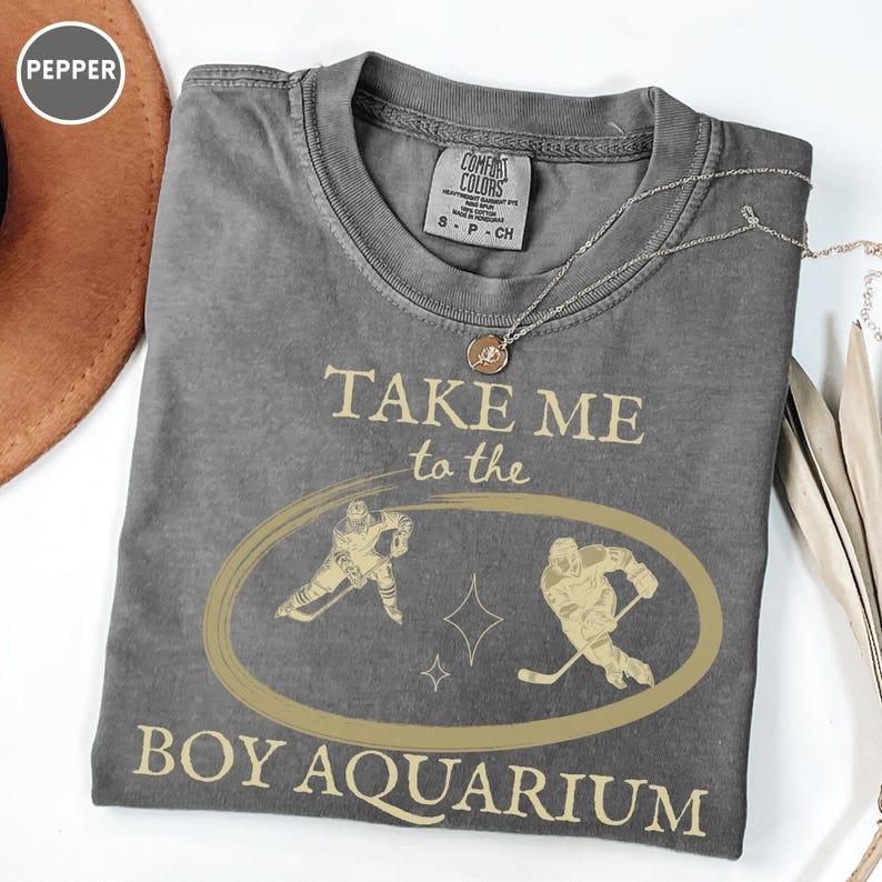 May include: A gray Comfort Colors t-shirt with the text "TAKE ME to the BOY AQUARIUM" in a gold color. The shirt features a gold oval with two hockey players and a star design. A silver necklace is draped across the shirt.