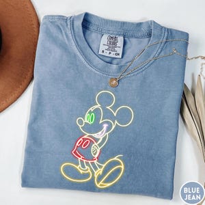 Op de afbeelding: Een blauw spijkerbroekkleurig T-shirt met een neon-stijl Mickey Mouse-afbeelding. De afbeelding heeft een gele omtrek met groene, rode en paarse accenten. Het shirt heeft een ronde hals en korte mouwen. Een gouden ketting ligt over het shirt.