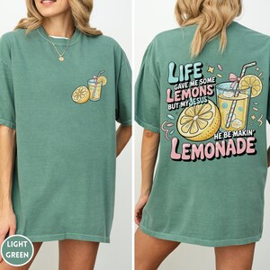 May include: Light green t-shirt with a front pocket graphic of a lemon and a drink. The back of the shirt features the text "LIFE GAVE ME SOME LEMONS BUT MY JESUS HE BE MAKIN' LEMONADE" with a lemon and drink graphic.