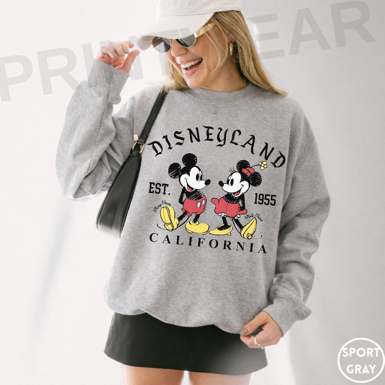 May include: Sport gray sweatshirt featuring Mickey and Minnie Mouse with the text "Disneyland Est. 1955 California". The graphic shows Mickey and Minnie Mouse in their classic outfits.