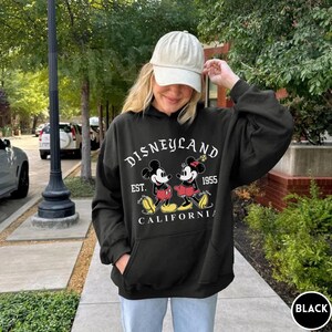 May include: Black hooded sweatshirt with a Disneyland graphic featuring Mickey and Minnie Mouse. The design includes the text "DISNEYLAND EST. 1955 CALIFORNIA" in white. The sweatshirt has a front pocket and a drawstring hood.