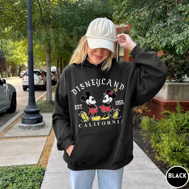 May include: Black hooded sweatshirt with a Disneyland graphic featuring Mickey and Minnie Mouse. The design includes the text "DISNEYLAND EST. 1955 CALIFORNIA" in white. The sweatshirt has a front pocket and a drawstring hood.