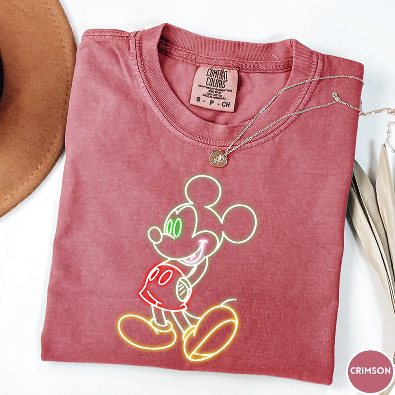 Op de afbeelding: Karmizijnrood T-shirt met een Mickey Mouse-ontwerp in neonstijl. De afbeelding bevat groene ogen, een rode broek en gele contouren. Een zilveren ketting met een hanger is over het shirt gedrapeerd. Het shirt heeft een Comfort Colors-label.