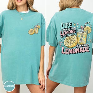 May include: Seafoam green t-shirt with a lemon and lemonade graphic. The front features a lemon and a glass of lemonade. The back of the shirt has the text "LIFE GAVE ME SOME LEMONS BUT MY JESUS HE BE MAKIN' LEMONADE."