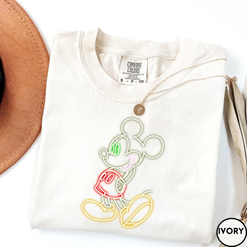 Op de afbeelding: Ivoor kleurig T-shirt met een Mickey Mouse-ontwerp in neonstijl. Het ontwerp bevat groene ogen, een roze mond, een rode broek en gele contouren. Het shirt heeft een Comfort Colors-label en een gouden ketting. Het woord "IVORY" staat in een cirkel.