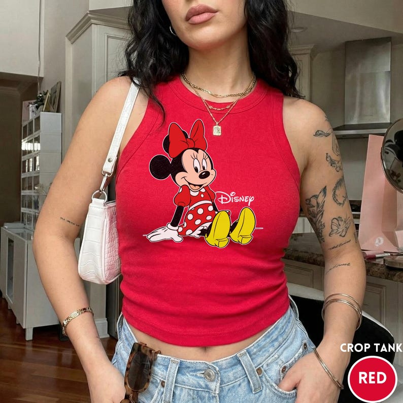 Puede incluir: Top corto rojo con un gr&aacute;fico de Minnie Mouse. El top sin mangas tiene un cuello redondo y una silueta ajustada. El dise&ntilde;o de Minnie Mouse incluye un lazo rojo, un vestido con lunares blancos y zapatos amarillos. La palabra "Disney" est&aacute; impresa debajo.
