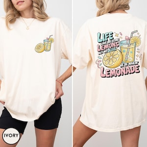 May include: Ivory-colored t-shirt with a graphic of a lemon and a glass of lemonade on the front. The back of the shirt features the text "Life gave me some lemons but my Jesus he be makin' lemonade" in pastel colors.