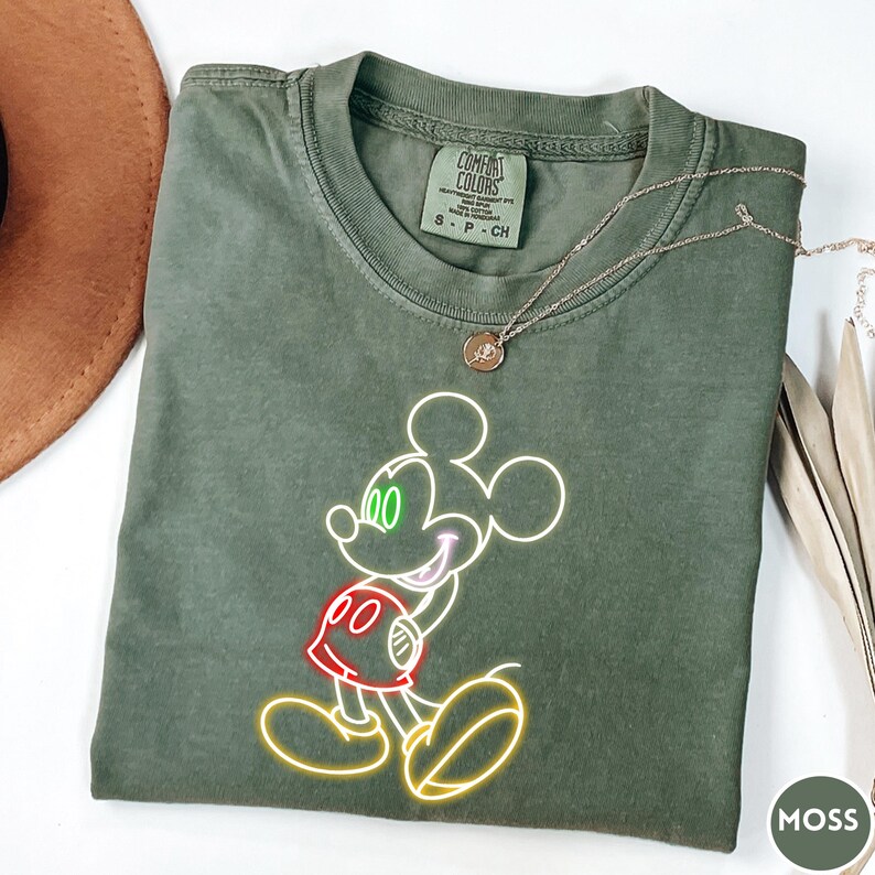 Op de afbeelding: Mosgroen Comfort Colors T-shirt met een neon-stijl Mickey Mouse ontwerp. De afbeelding bevat gele, rode en groene accenten. Een zilveren ketting met een hanger is over het shirt gedrapeerd.
