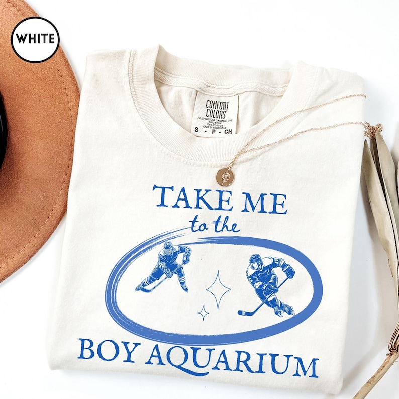 May include: Cream-colored t-shirt with the text "TAKE ME to the BOY AQUARIUM" in blue. The design includes two hockey players inside a blue oval, with a star graphic. A gold necklace is draped across the shirt.
